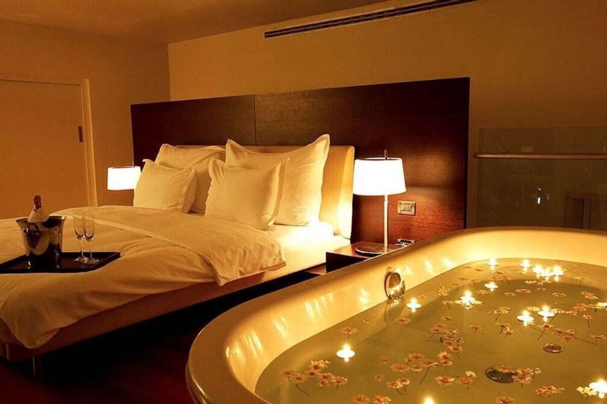 A hotel room with a bed, bedside lamps, a tray with a bottle and glasses, and a bathtub with candles and flowers.