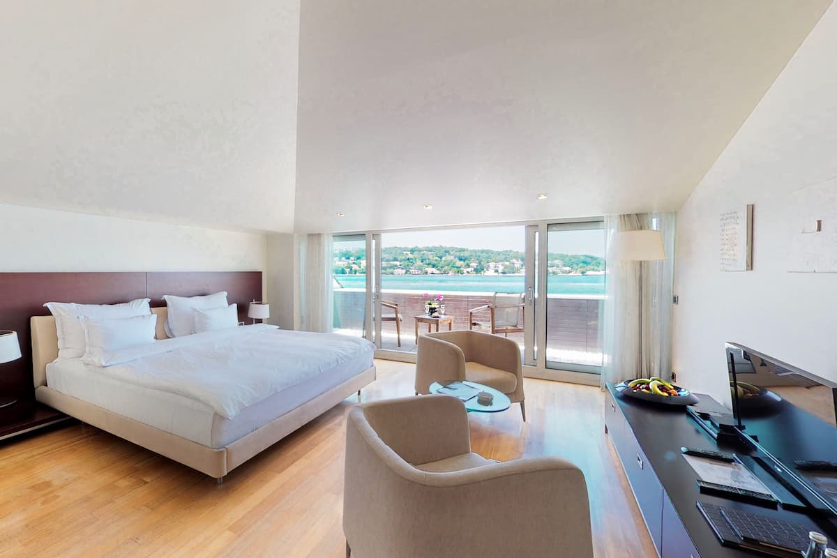 A modern hotel room with a large bed, a desk with a computer, and a balcony with a view of the sea.