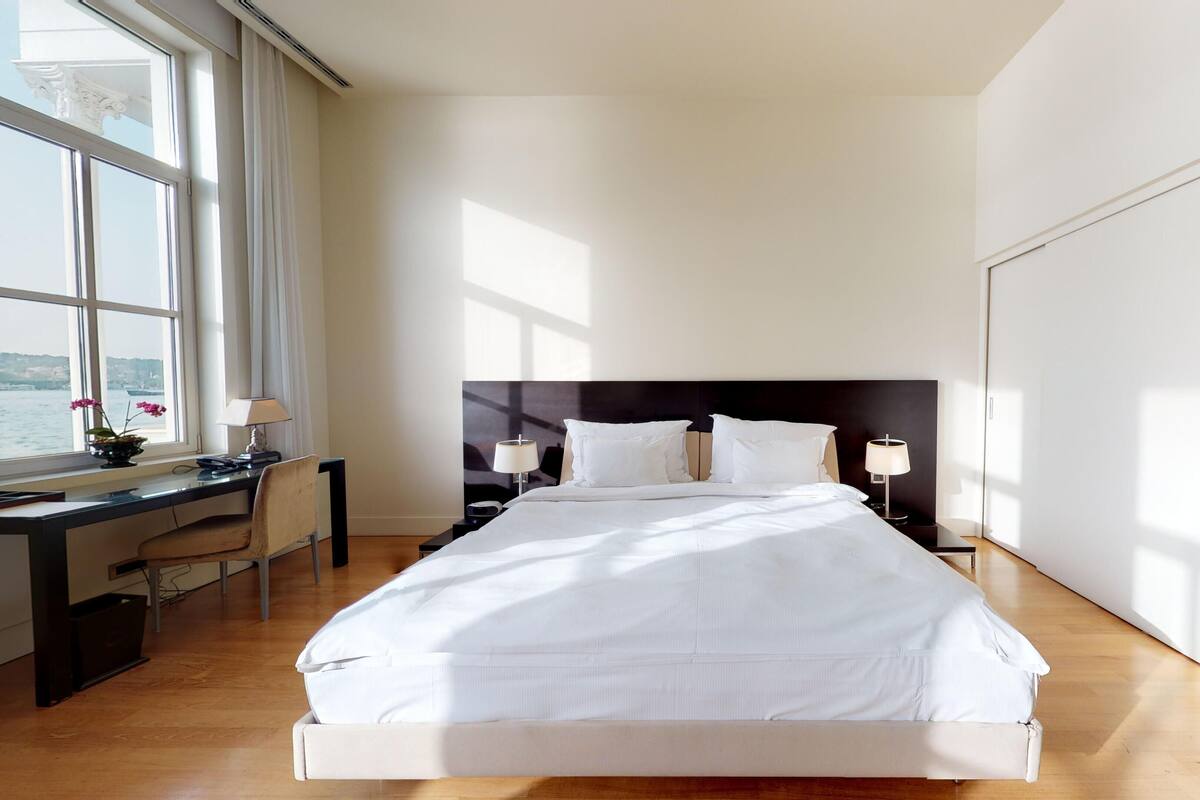A modern hotel room with a large bed, a desk with a chair, a nightstand with lamps, and a view of the water through the window.