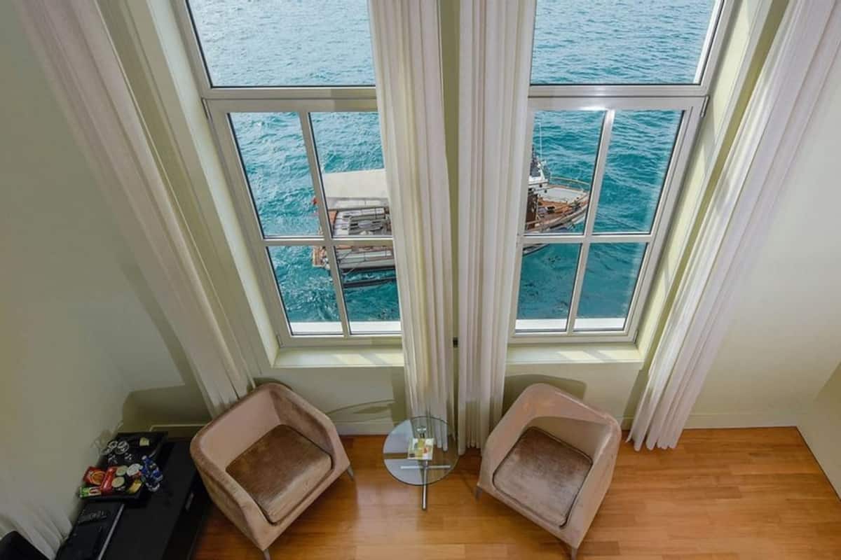 A room with a large window overlooking a body of water, two cushioned chairs, a small table, and a wooden floor.