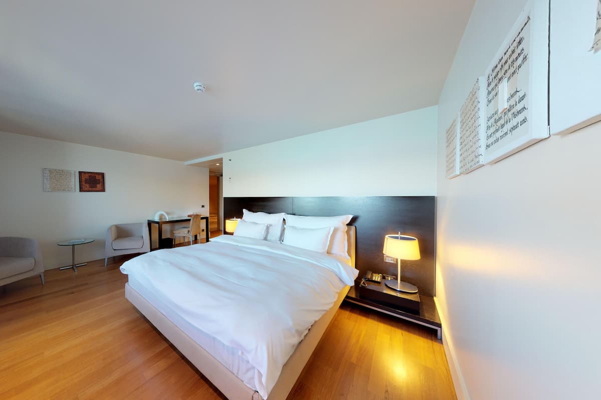 A hotel room with a large bed, a desk, two chairs, a nightstand with a lamp, and a wall-mounted artwork.