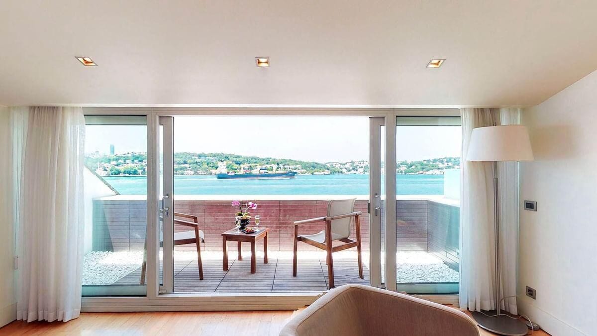 Ein Zimmer mit Balkon, einem Tisch mit Stühlen, einer Lampe und Blick auf das Meer sowie entfernte Gebäude.
