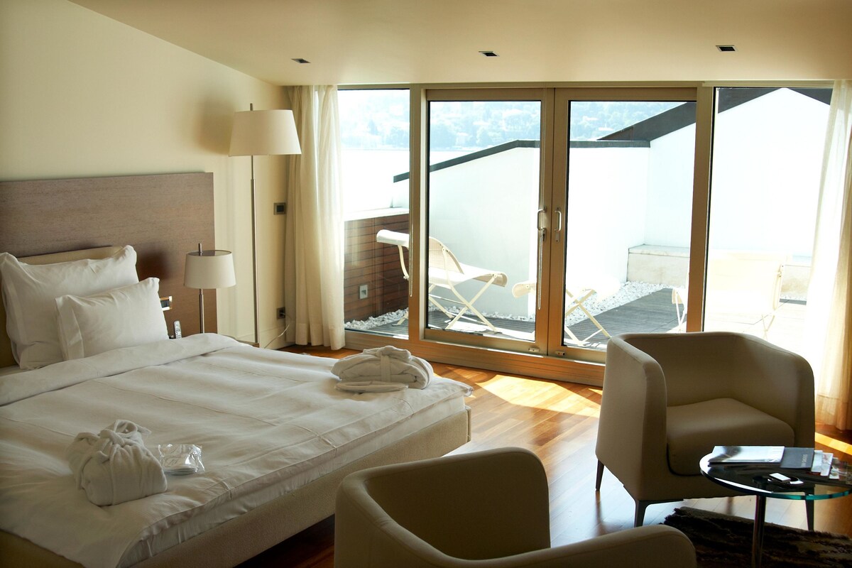 A hotel room with a large bed, a sitting area with a chair and a table, and a balcony with a chair and a table.