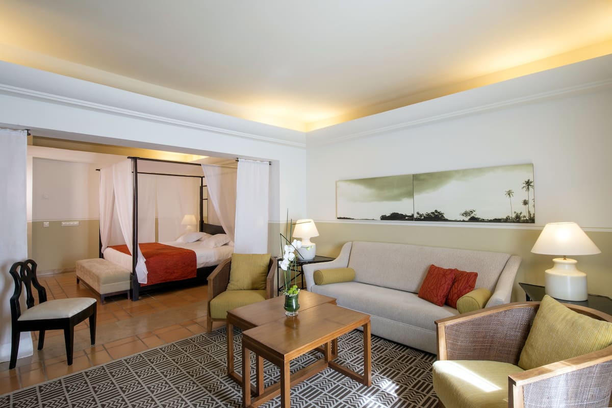 Une chambre d’hôtel avec un lit, un canapé, une table basse et une chaise. Il y a un tableau au mur et une lampe sur la table de chevet.