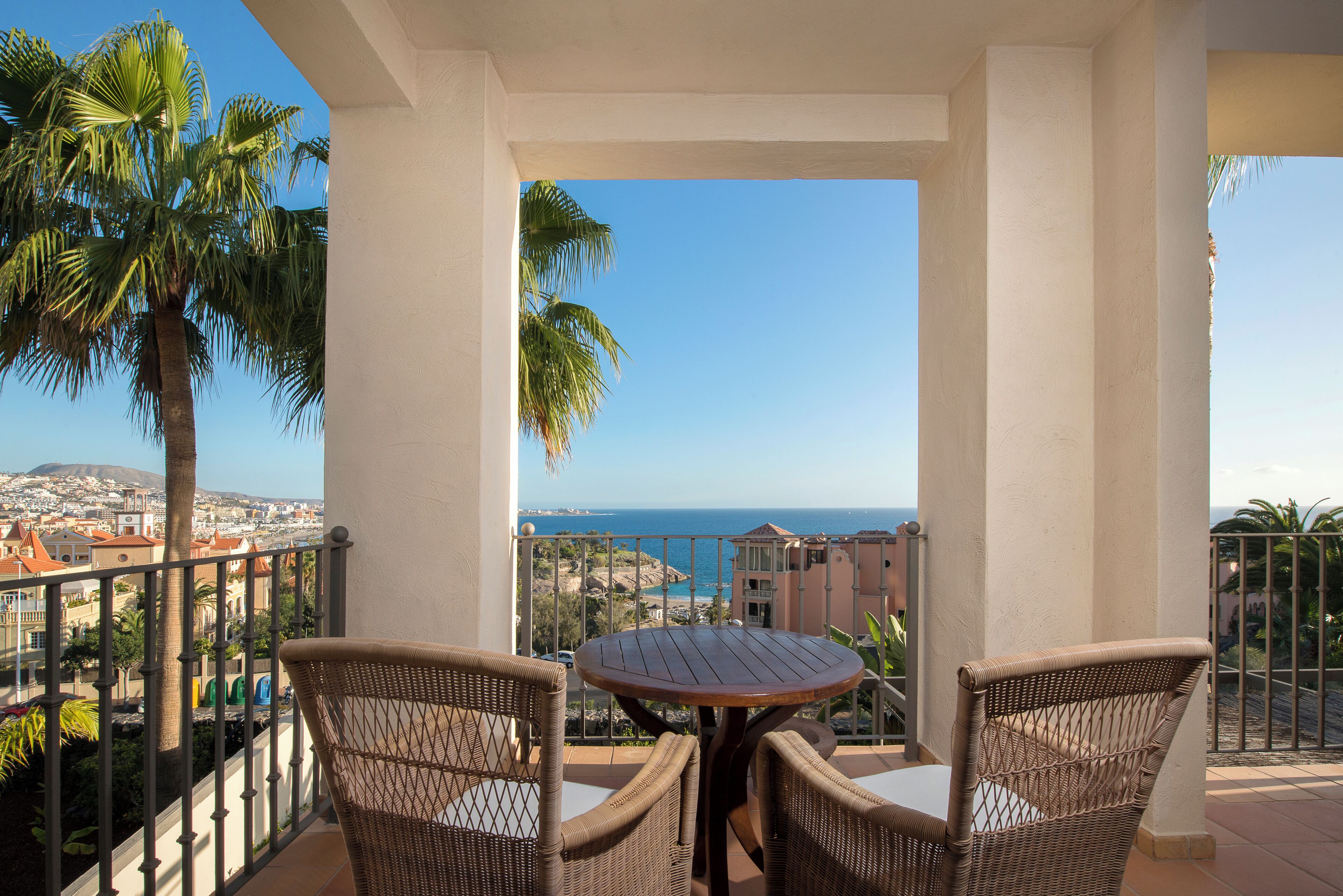 A balcony with wicker chairs and a table, overlooking a coastal cityscape and the sea.