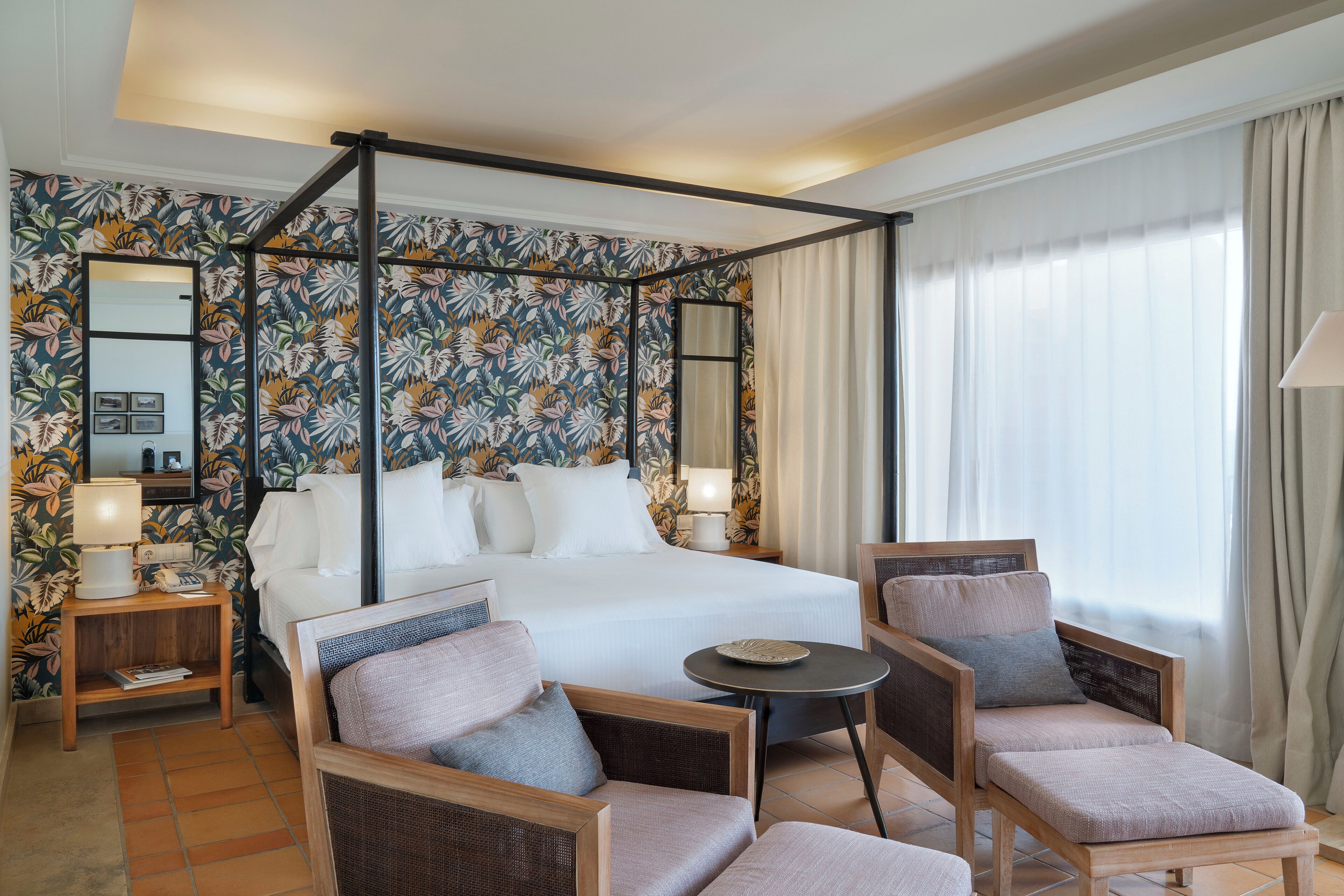 Una cama con dosel, papel tapiz floral, un rincón de estar con dos sillones y una mesita, y un ventanal con cortinas.