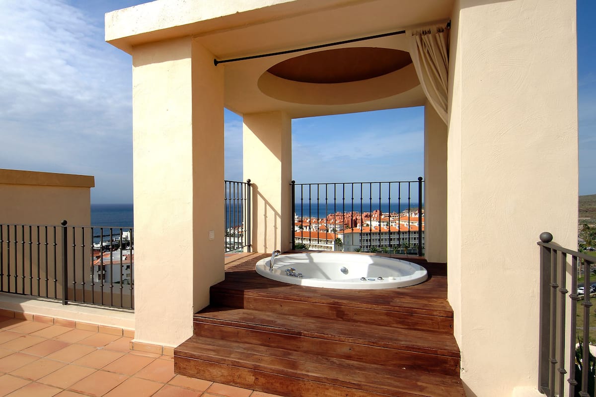 Un balcon doté d’un jacuzzi, d’un escalier en bois et offrant une vue sur la mer et les bâtiments.