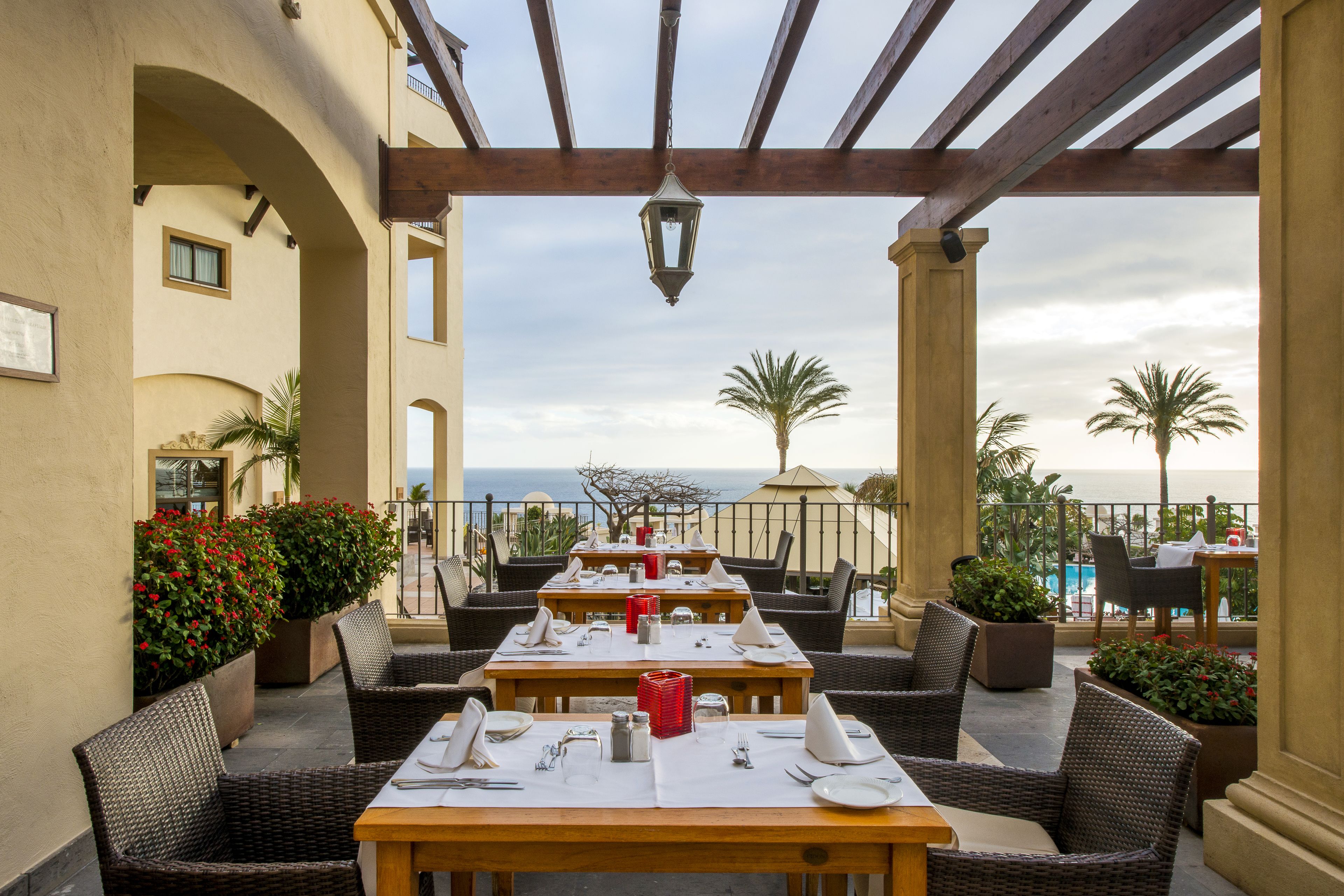 An outdoor dining area with tables set for two, overlooking a pool and the ocean.
