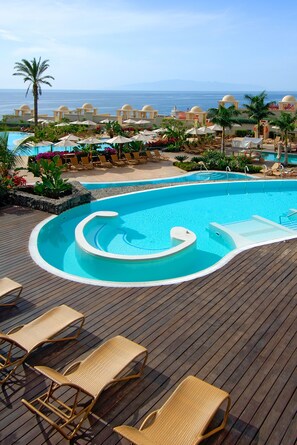 Zona de piscina con sillones de descanso y sombrillas, una terraza de madera y vista al océano.
