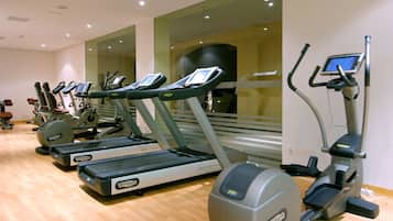 A row of cardio machines in a gym with a wooden floor and large windows.