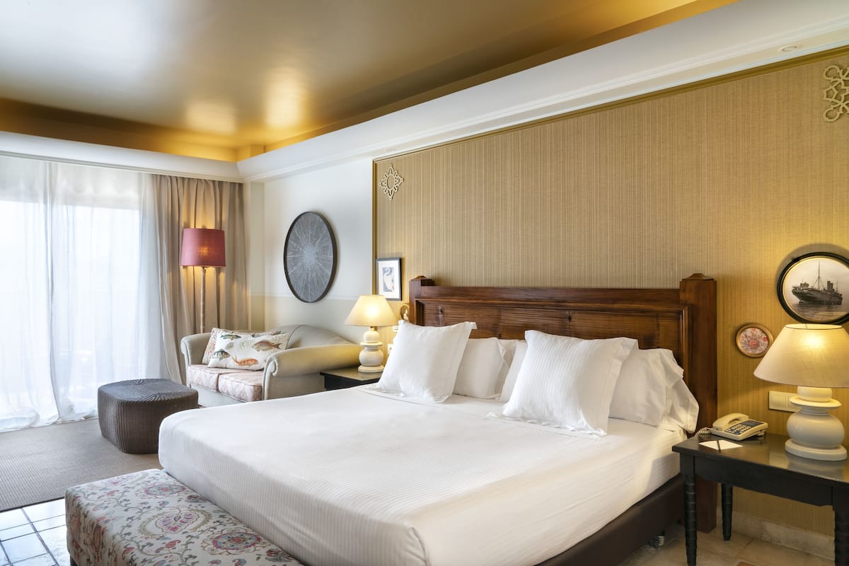 Une chambre d’hôtel avec un grand lit, une table de chevet avec une lampe, une chaise et une fenêtre avec des rideaux.