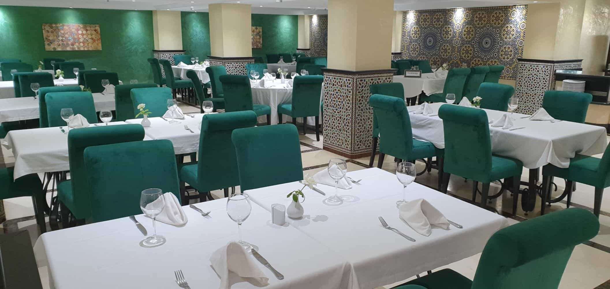 A restaurant with multiple tables, each set with white tablecloths, green chairs, and wine glasses.