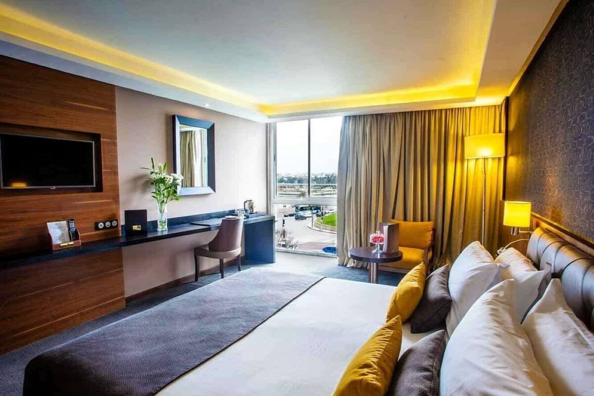 A hotel room with a large bed, a desk, a chair, a TV, and a view of the city.