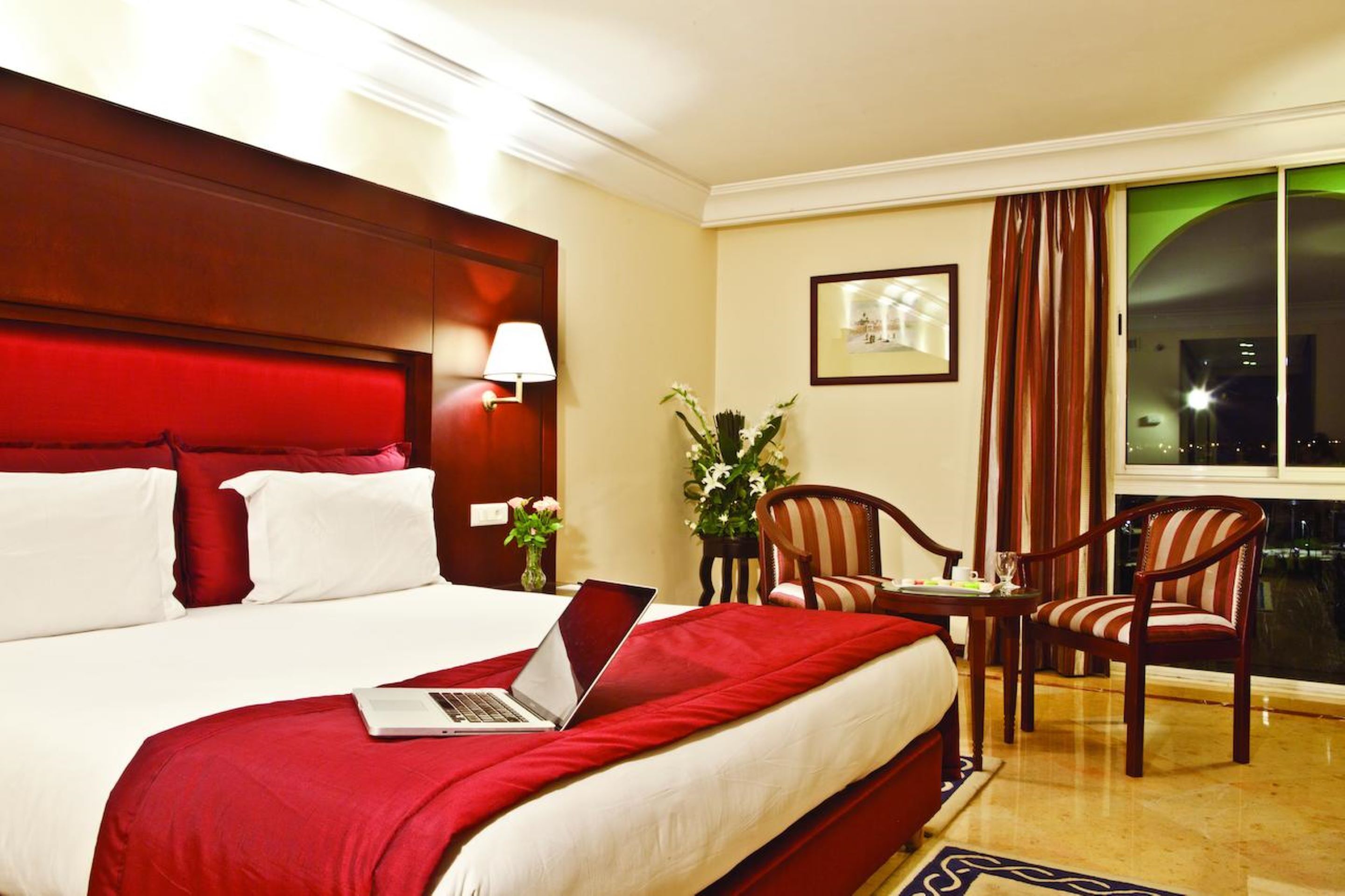 A hotel room with a large bed, a laptop on the bed, a desk with two chairs, a window with curtains, and a framed picture on the wall.