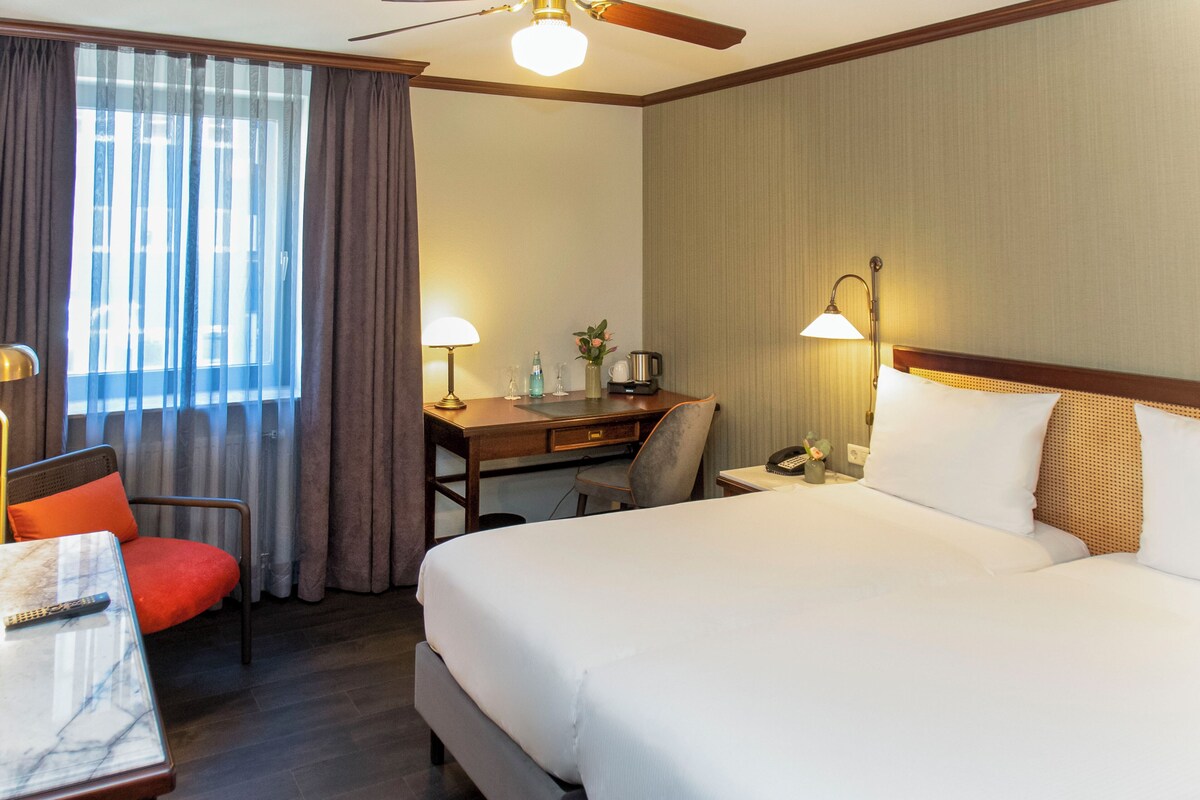 A hotel room with a large bed, a desk with a chair, a desk lamp, a telephone, and a window with curtains.