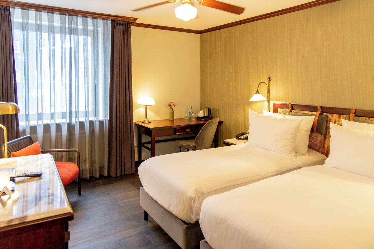 A hotel room with two beds, a desk, a chair, a lamp, and a window with curtains.