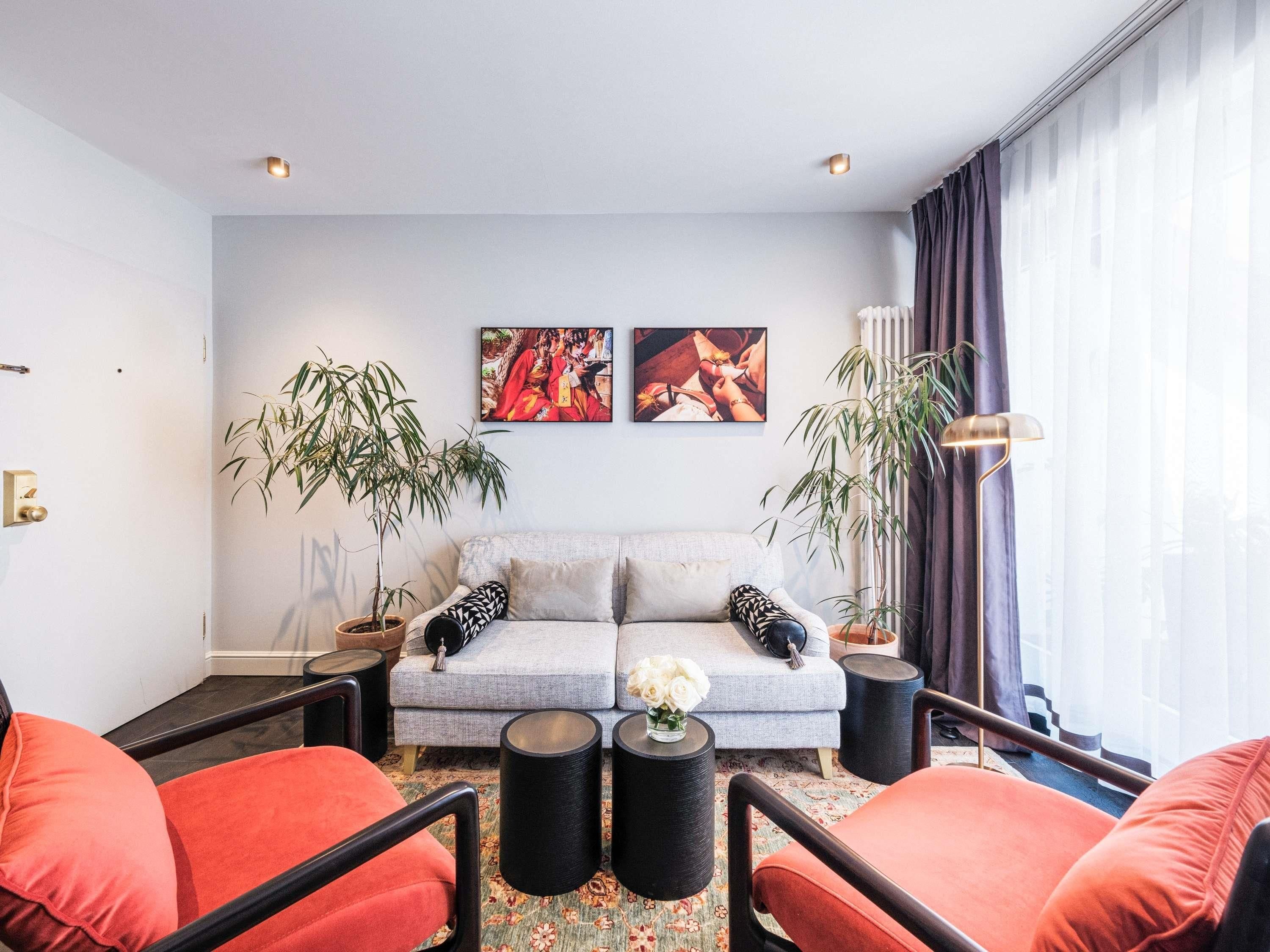 Ein modernes Wohnzimmer mit einem grauen Sofa, roten Sessel, zwei schwarzen Beistelltischen und zwei großen Kübelpflanzen.