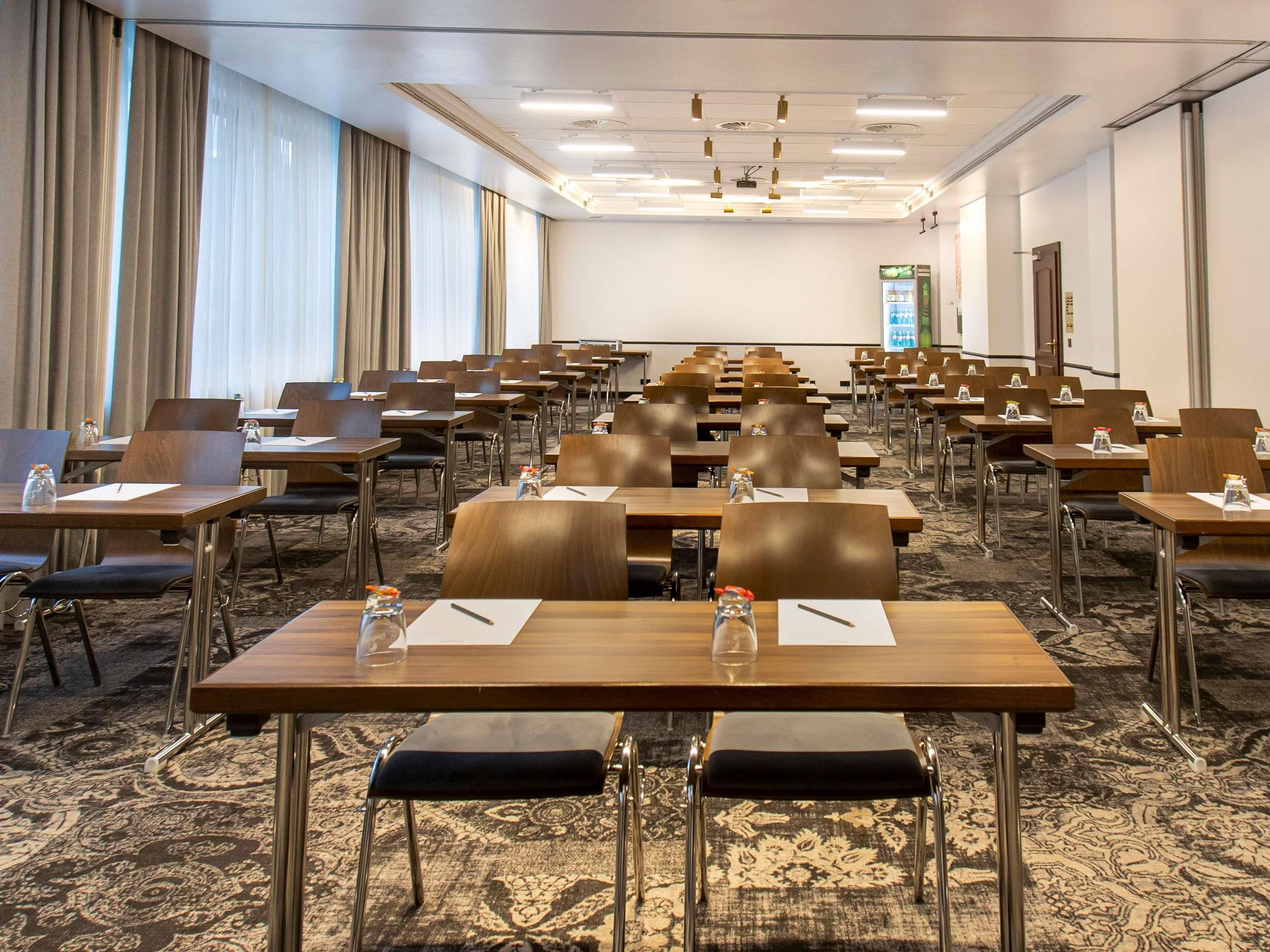 Une salle de conférence avec des rangées de tables et de chaises, un écran de projection et un sol recouvert de moquette.