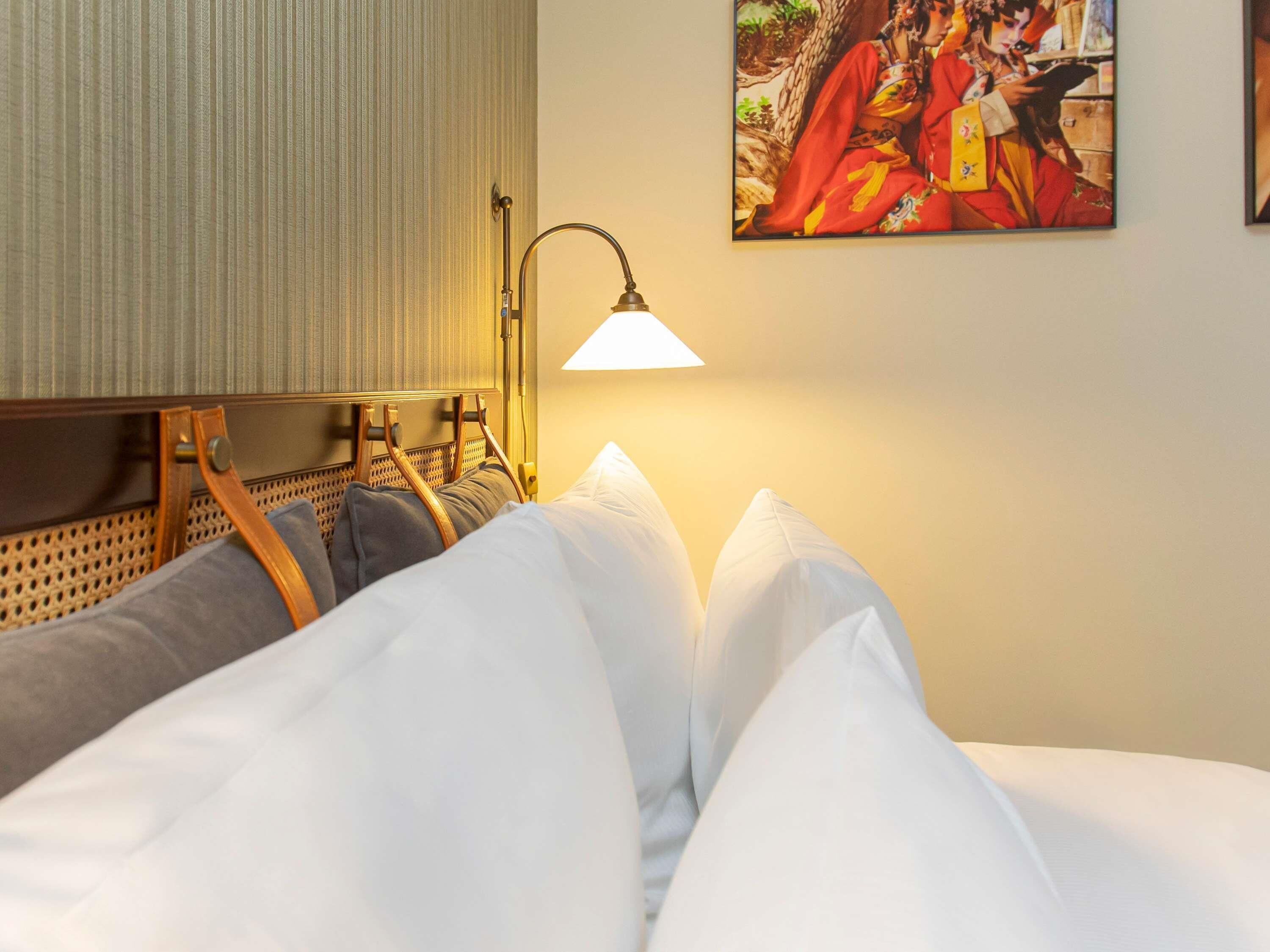 A hotel room with a bed, pillows, a headboard, a lamp, and a painting on the wall.