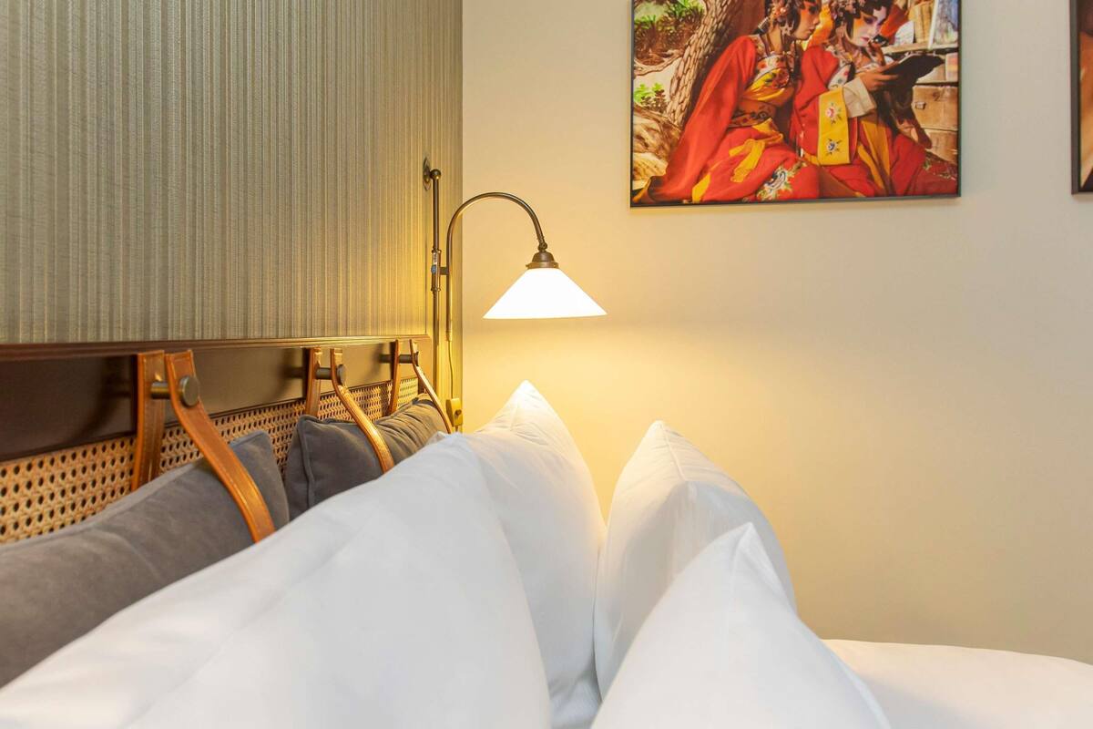 A hotel room with a bed, pillows, a headboard, a lamp, and a painting on the wall.