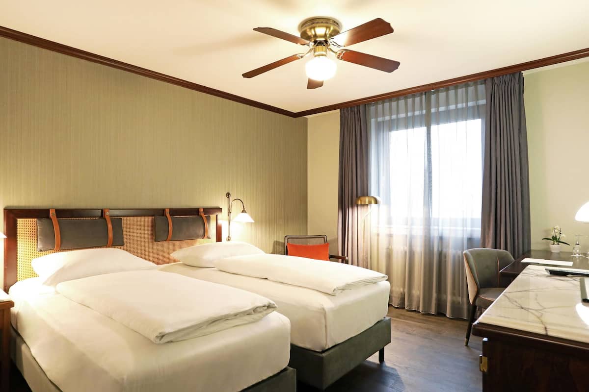 Une chambre d’hôtel avec deux lits, un bureau, une chaise, un ventilateur de plafond et une fenêtre avec des rideaux.