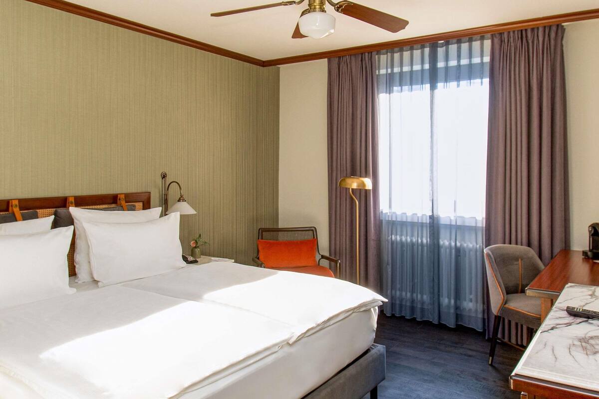 Une chambre d’hôtel avec un grand lit, un bureau, une chaise, une lampe et un ventilateur de plafond.