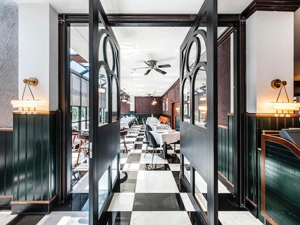 Un couloir au sol carrelé mène à une salle à manger avec des tables et des chaises.