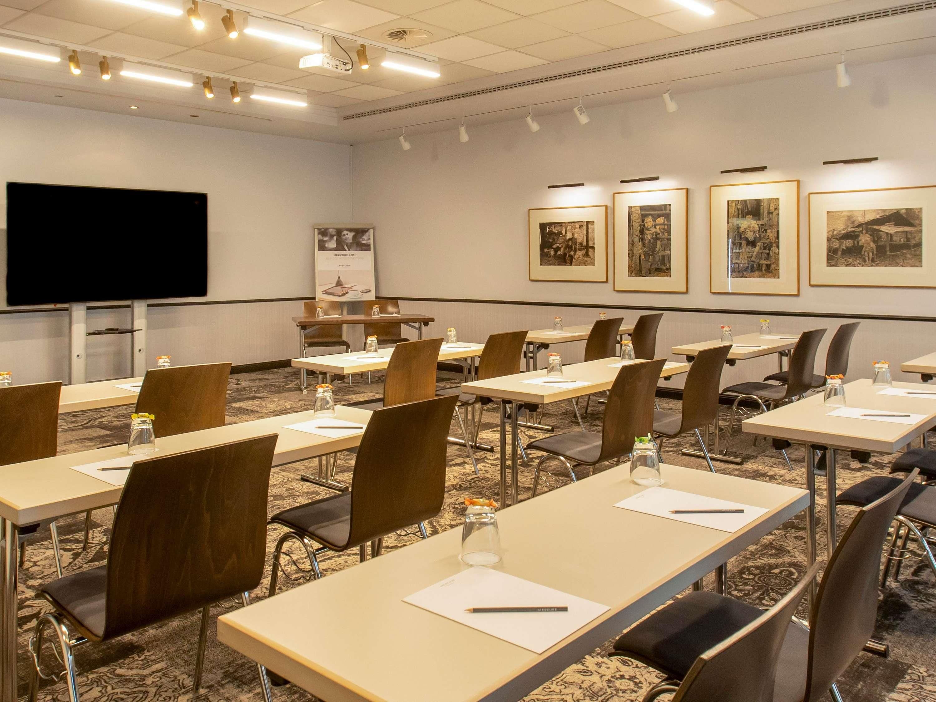 A conference room with long tables, chairs, a projector screen, and framed pictures on the wall.