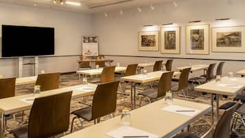 A conference room with long tables, chairs, a projector screen, and framed pictures on the wall.