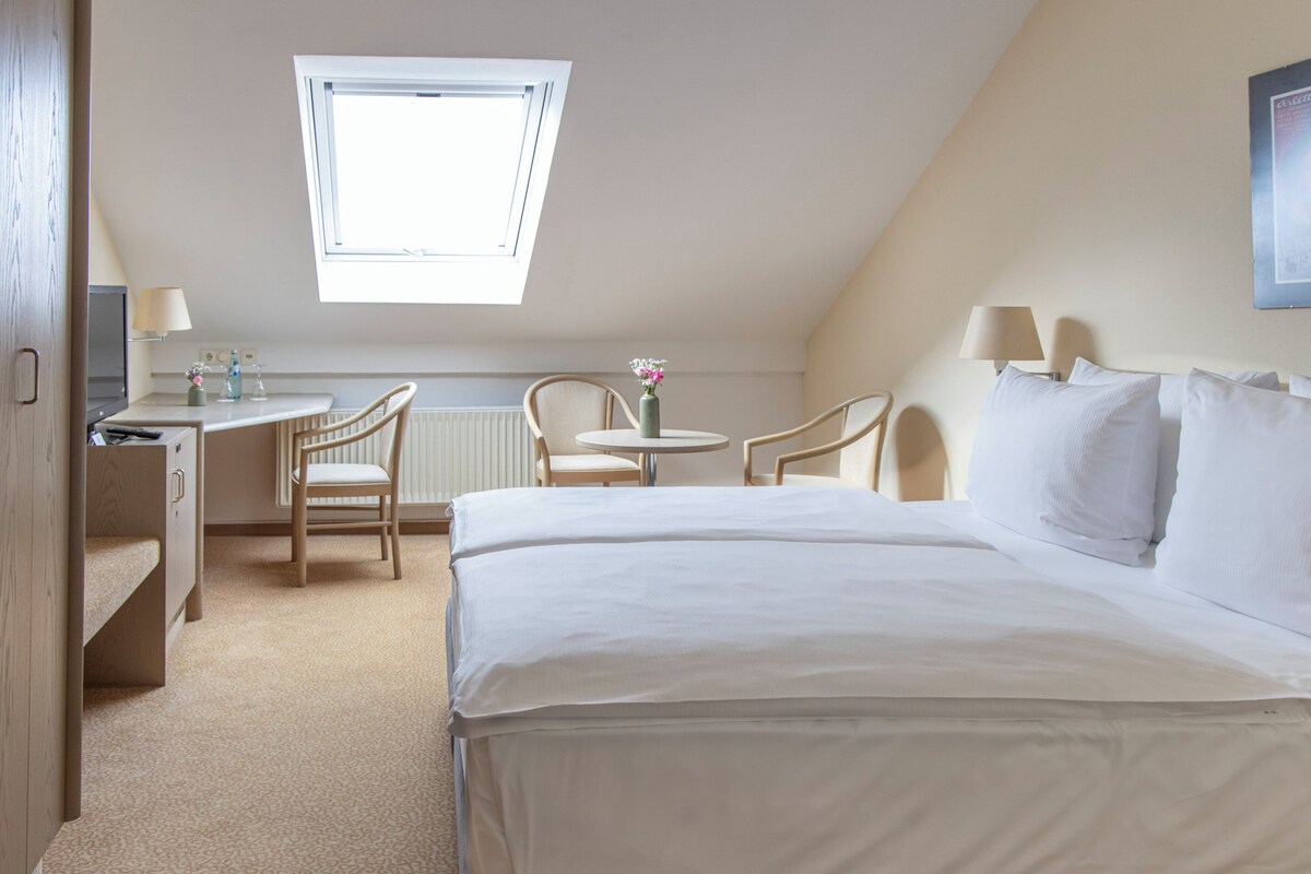 A hotel room with a large bed, a desk, two chairs, a small table with a vase, and a window providing natural light.
