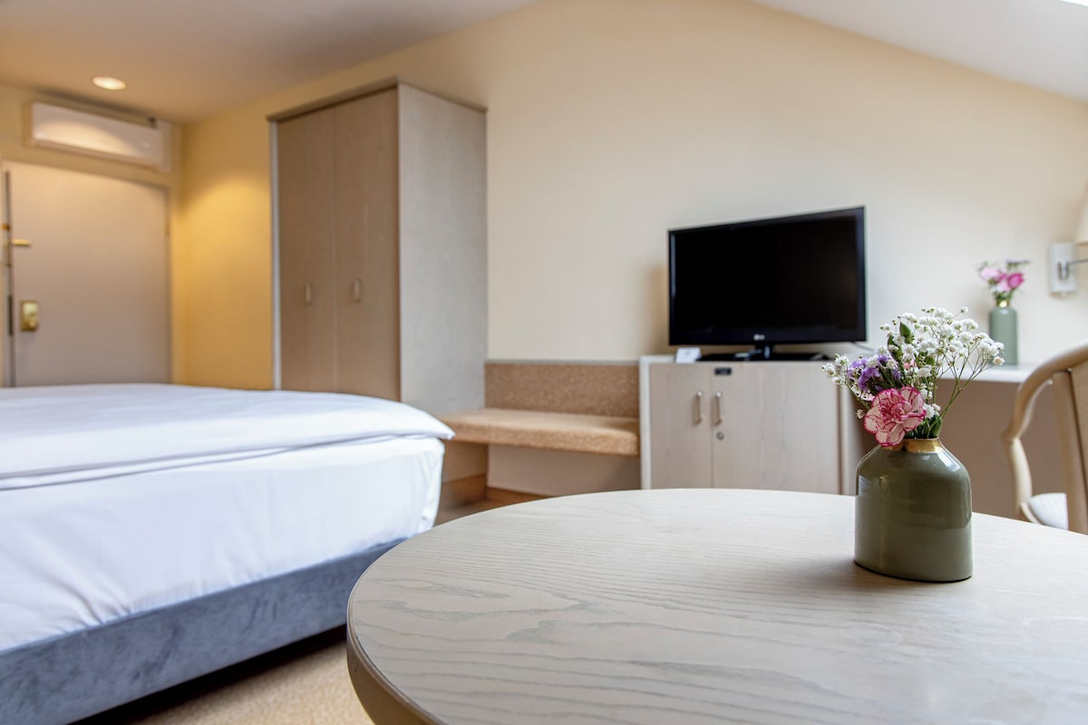 A hotel room with a bed, a television on a stand, a small table with a vase of flowers, and a wardrobe.
