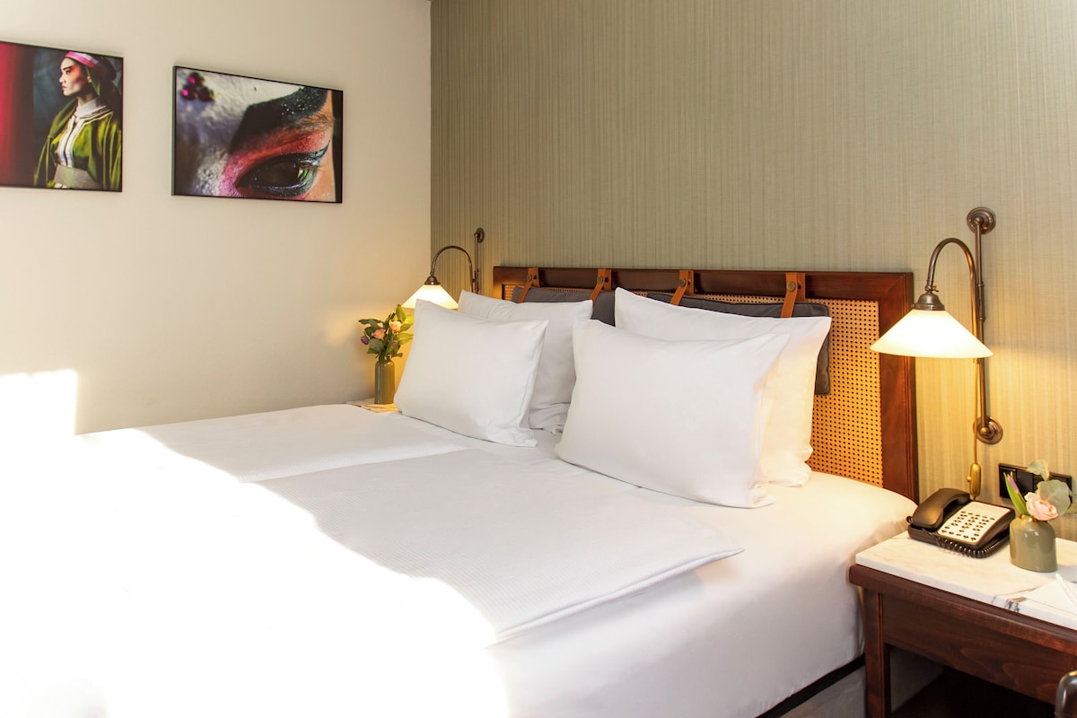 A neatly made bed with white linens, two wall-mounted lamps, a bedside telephone, and a vase with flowers.