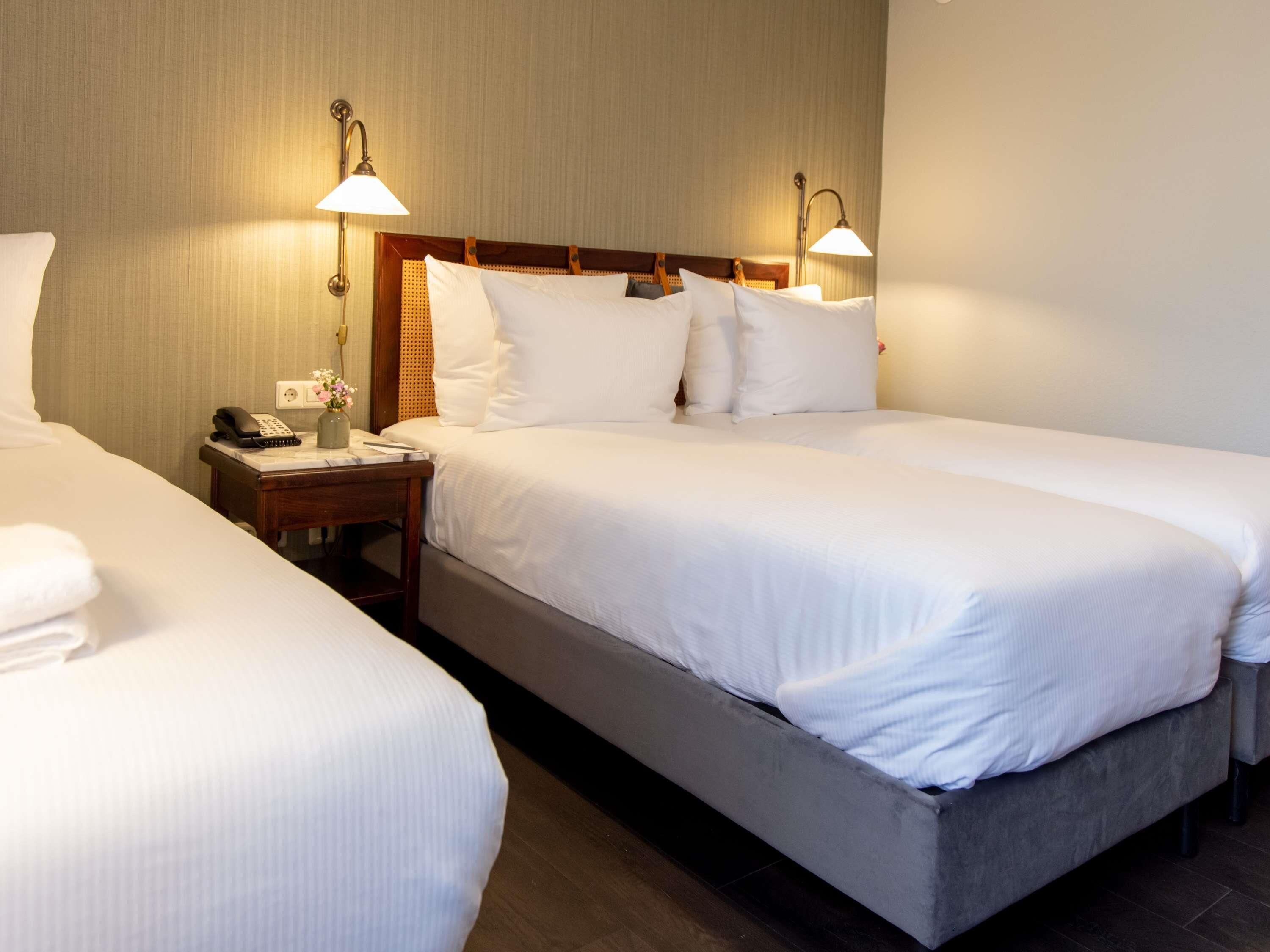A hotel room with two single beds, a bedside table with a lamp, a phone, and a vase with flowers.