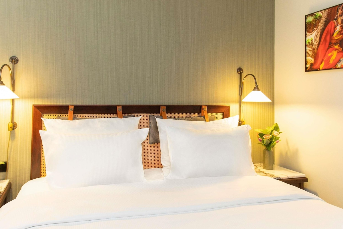 A hotel room with a bed, two bedside lamps, a nightstand with a vase of flowers, and a painting on the wall.