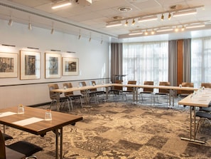 A conference room with a long table, chairs, framed pictures on the wall, and large windows with curtains.
