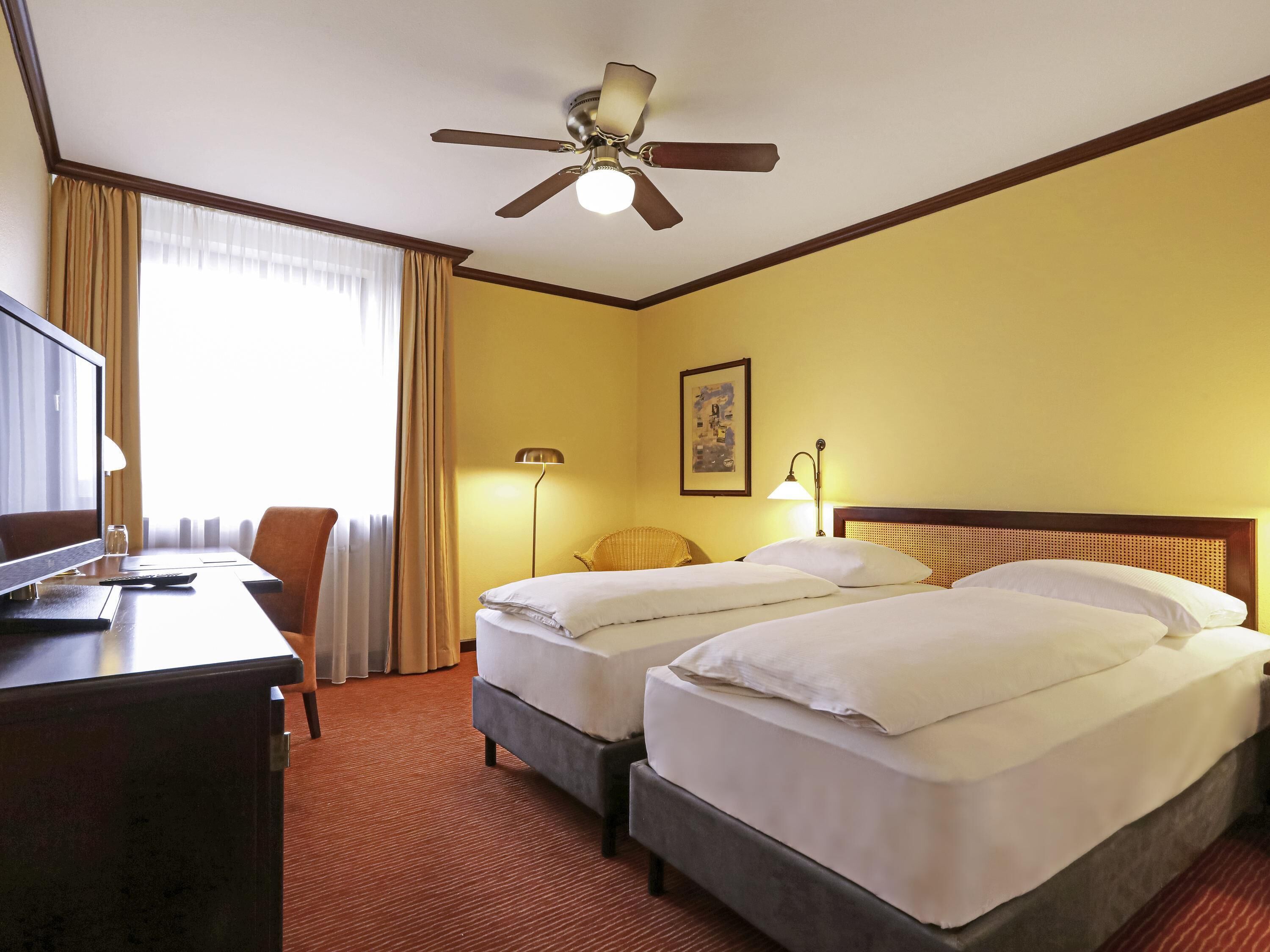 A hotel room with two beds, a desk with a TV, a ceiling fan, and a window with curtains.