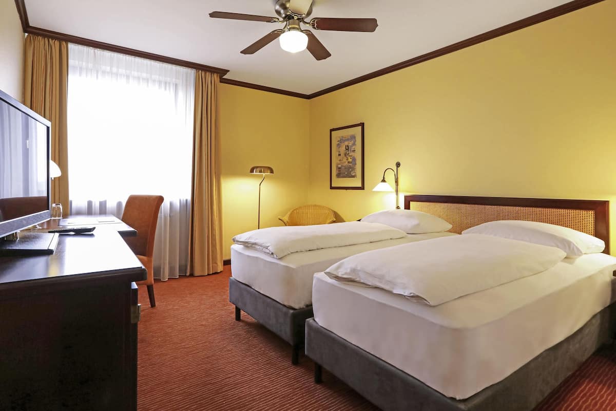 A hotel room with two beds, a desk with a TV, a ceiling fan, and a window with curtains.