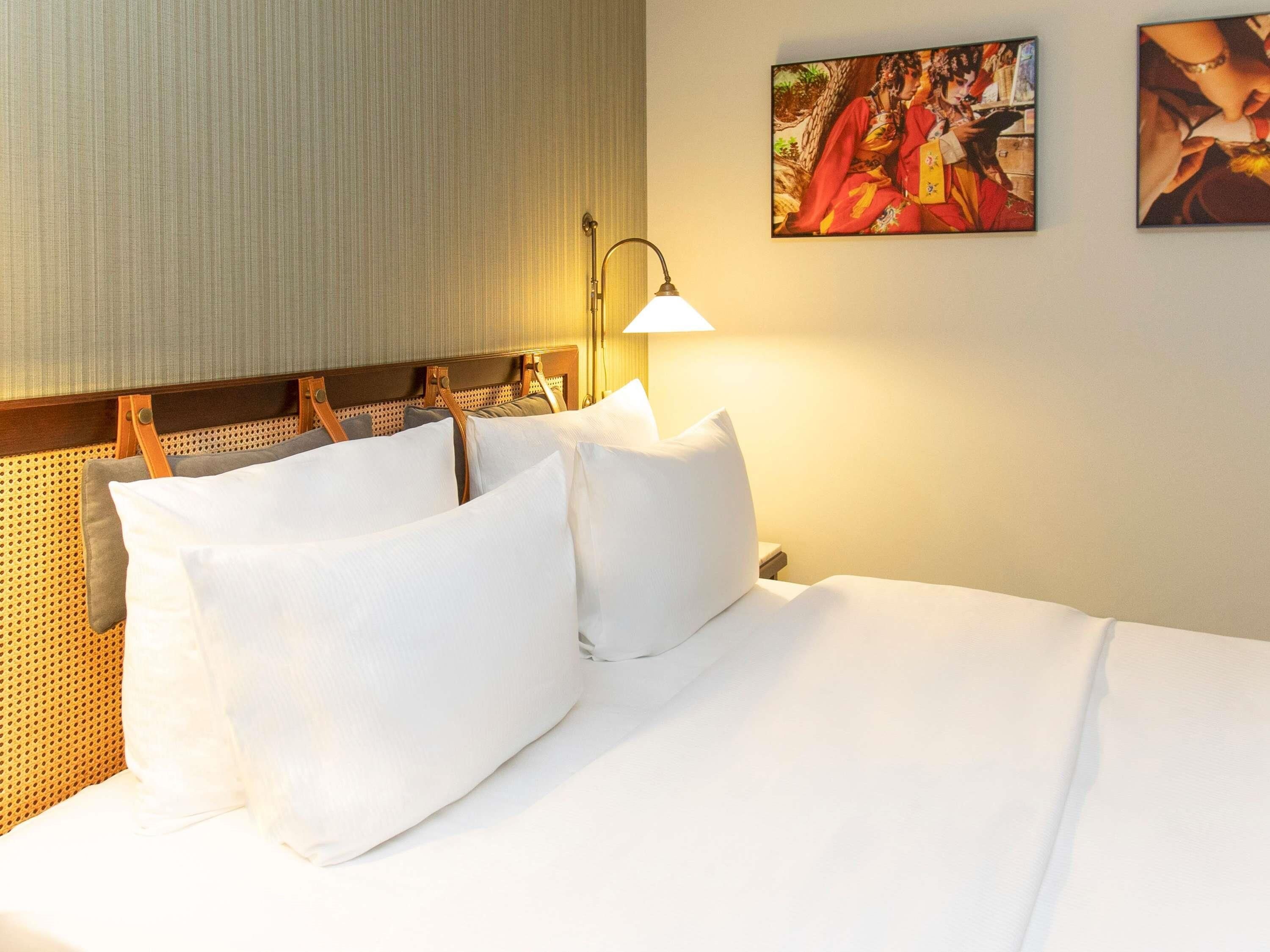 Ein Hotelzimmer mit einem Bett, Kissen, einem Kopfteil, einer Wandleuchte und zwei gerahmten Bildern.