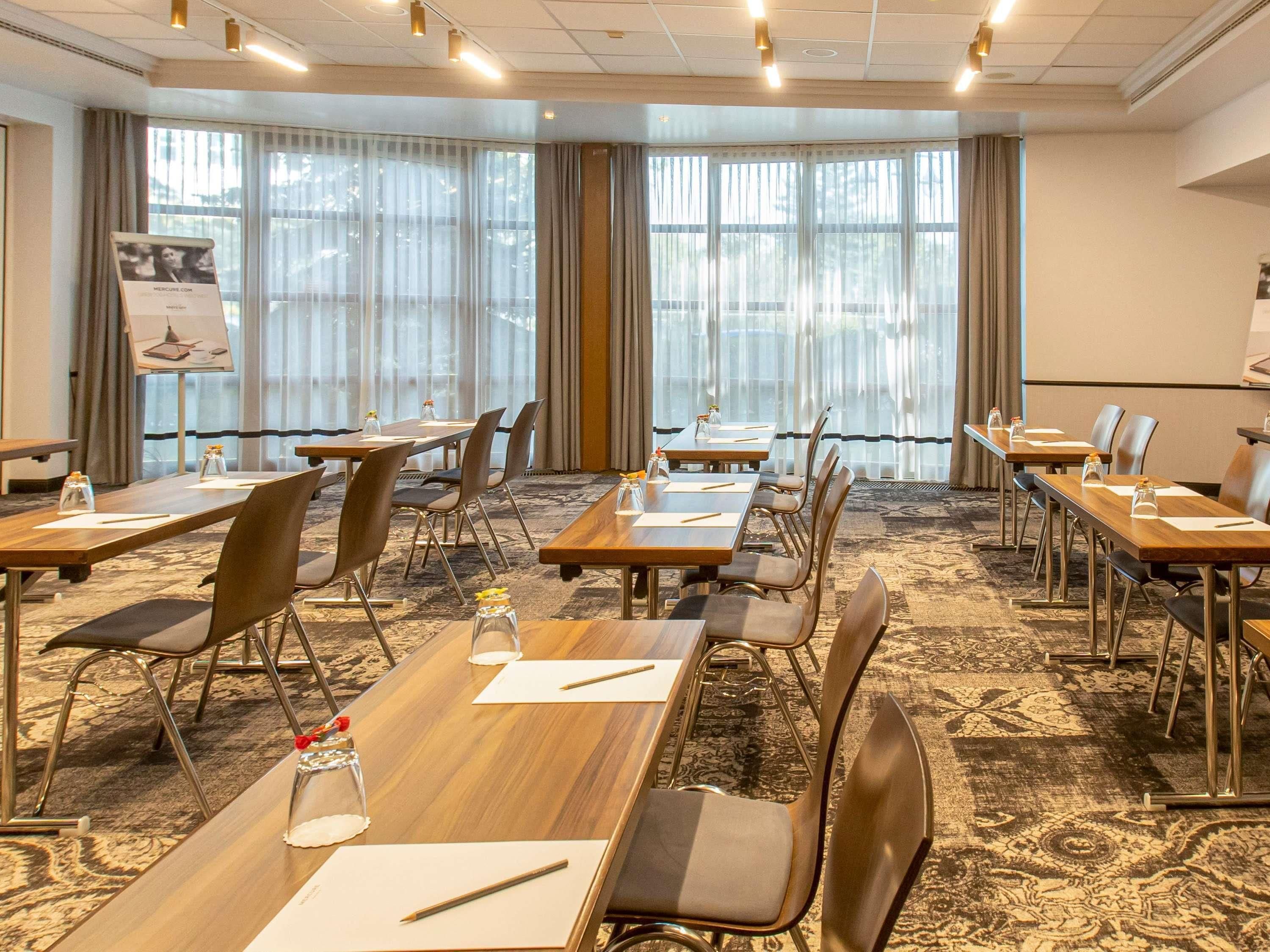 Ein Konferenzraum mit mehreren Tischen und Stühlen, großen Fenstern mit Vorhängen und einer Projektionsleinwand an der Wand.