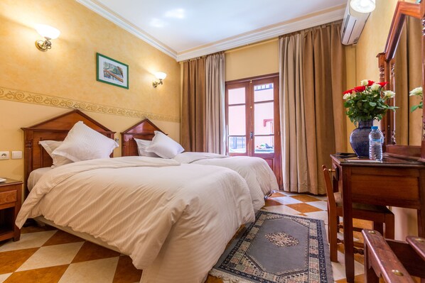 A hotel room with two beds, a wooden headboard, a nightstand, a desk with a vase of flowers, and a patterned rug. - Oudaya Hotel & Spa (Marrakech)