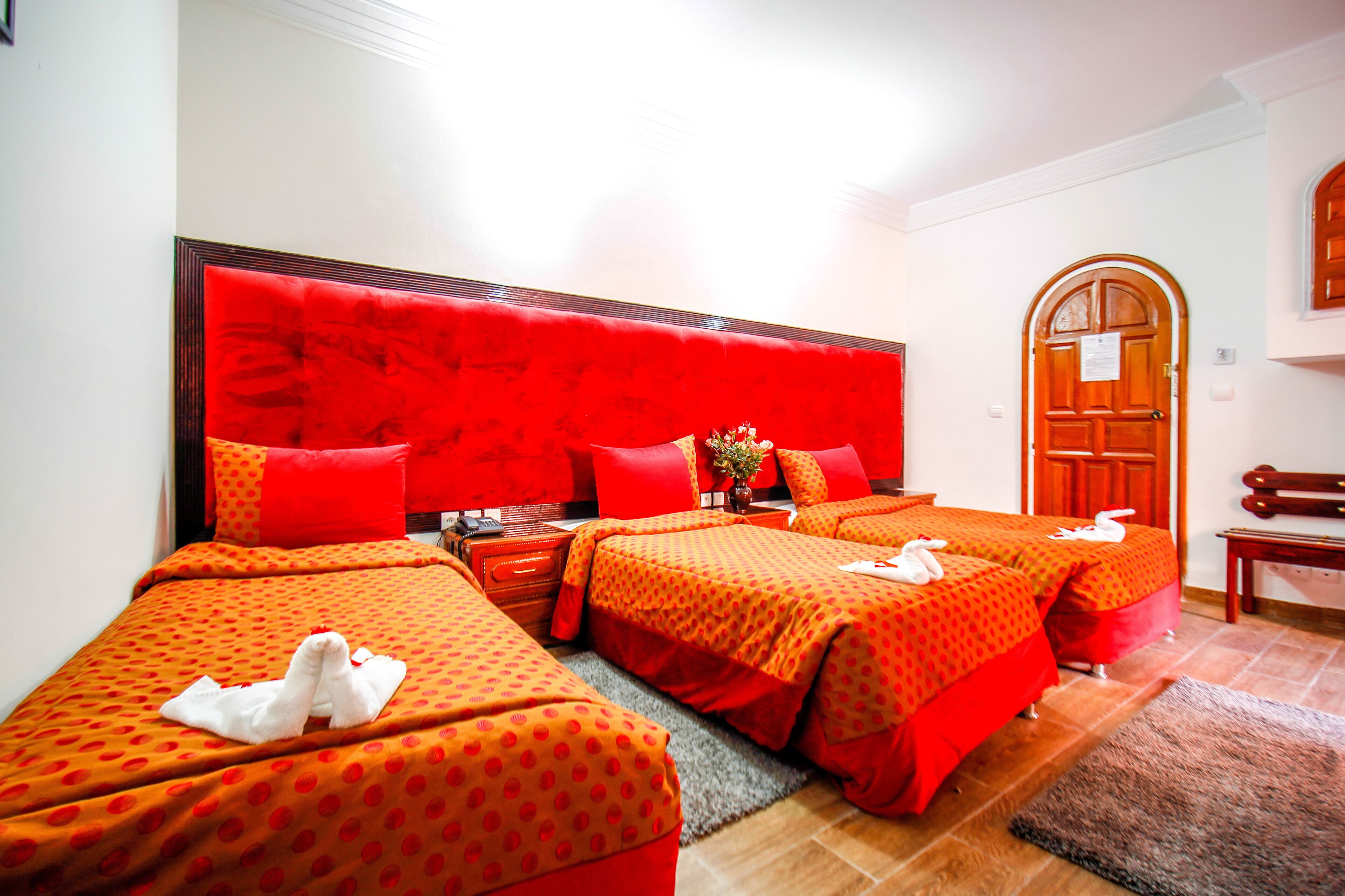 A hotel room with three beds, each with red bedding and white towels. There is a wooden door and a bench in the corner.