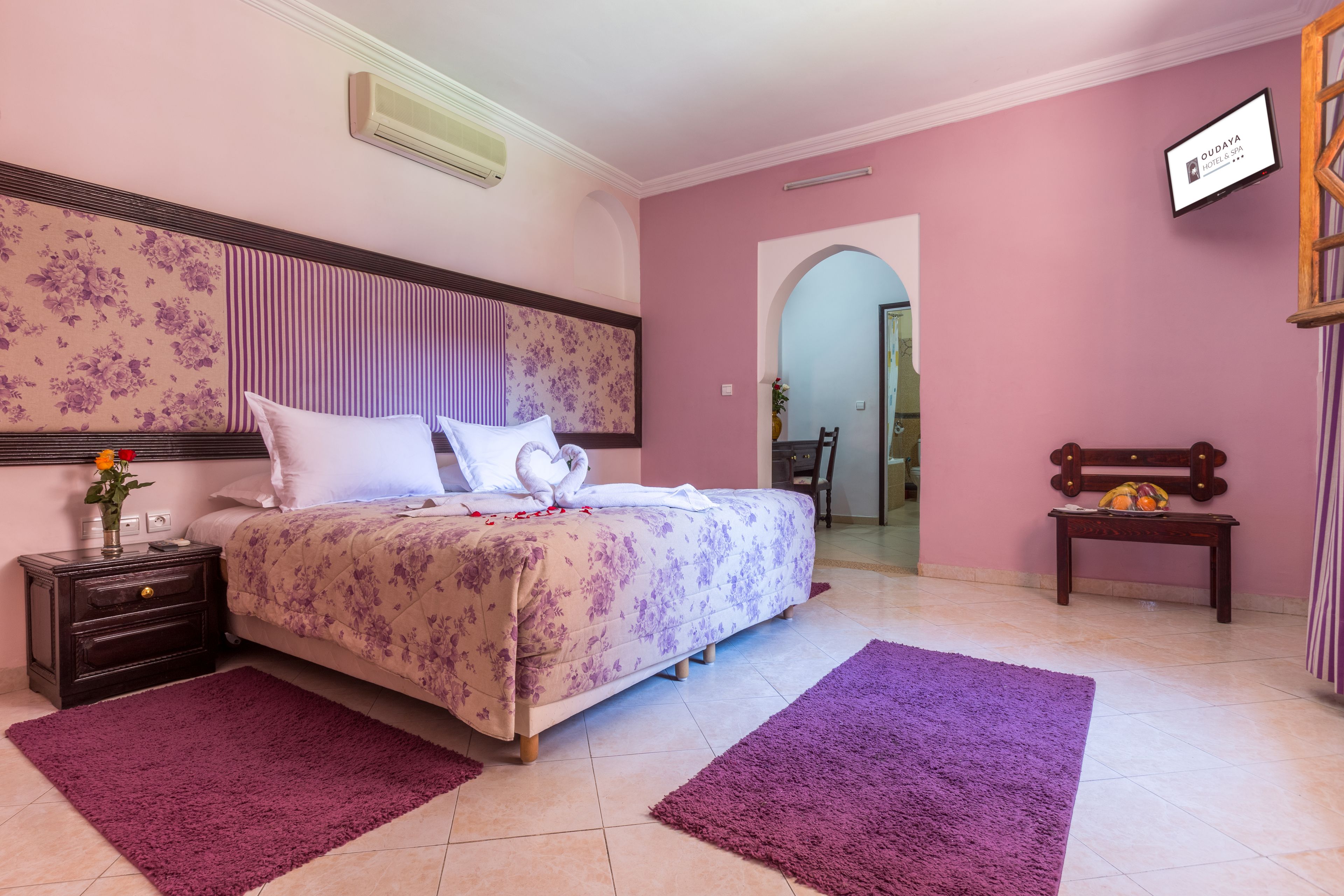 Une chambre à coucher avec une couvre-lit à motifs floraux, deux tapis violets, une table de chevet en bois ornée d’un vase de fleurs et un banc en bois avec un coussin.