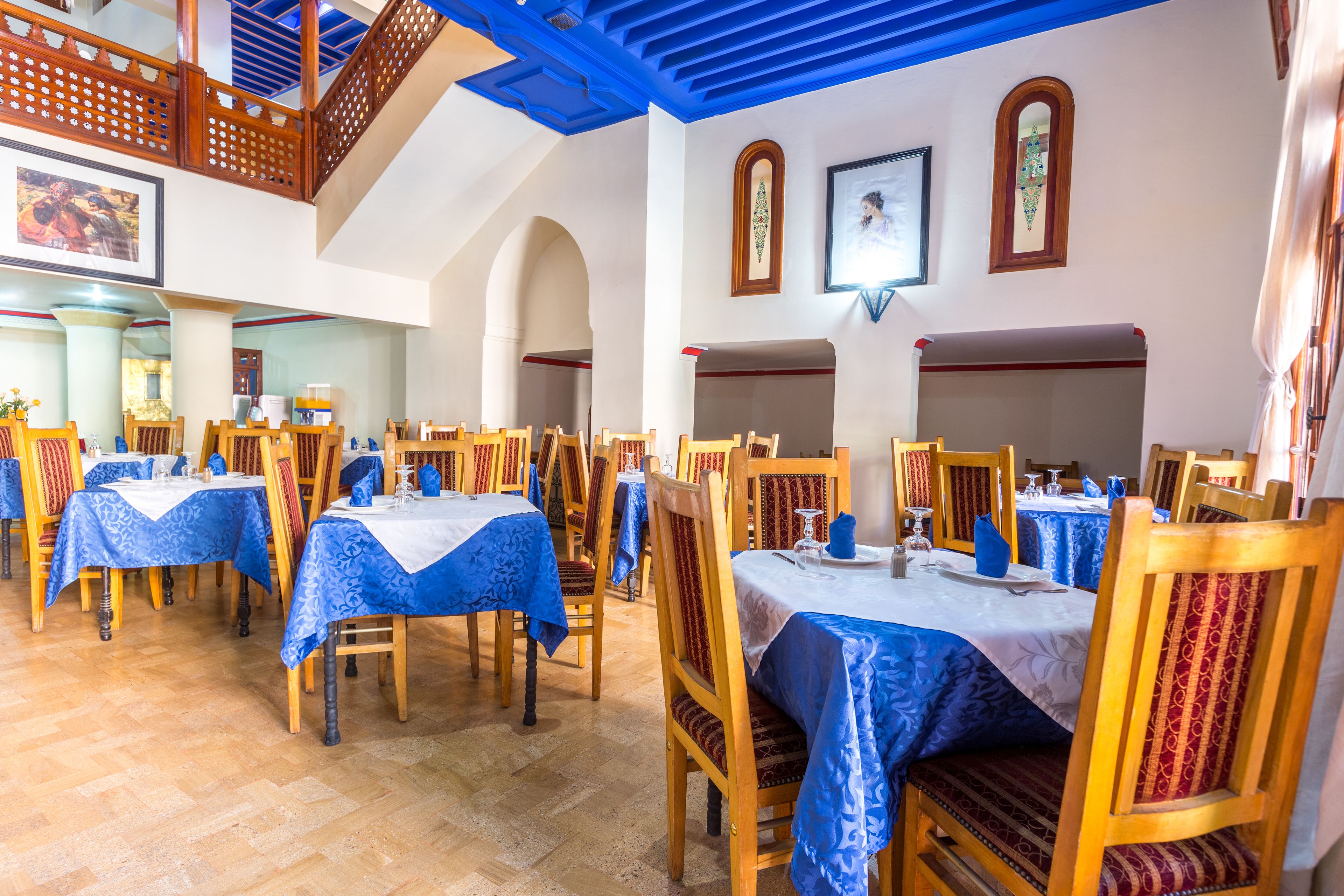 A dining area with multiple tables and chairs, blue tablecloths, wooden chairs, and a staircase with blue accents.