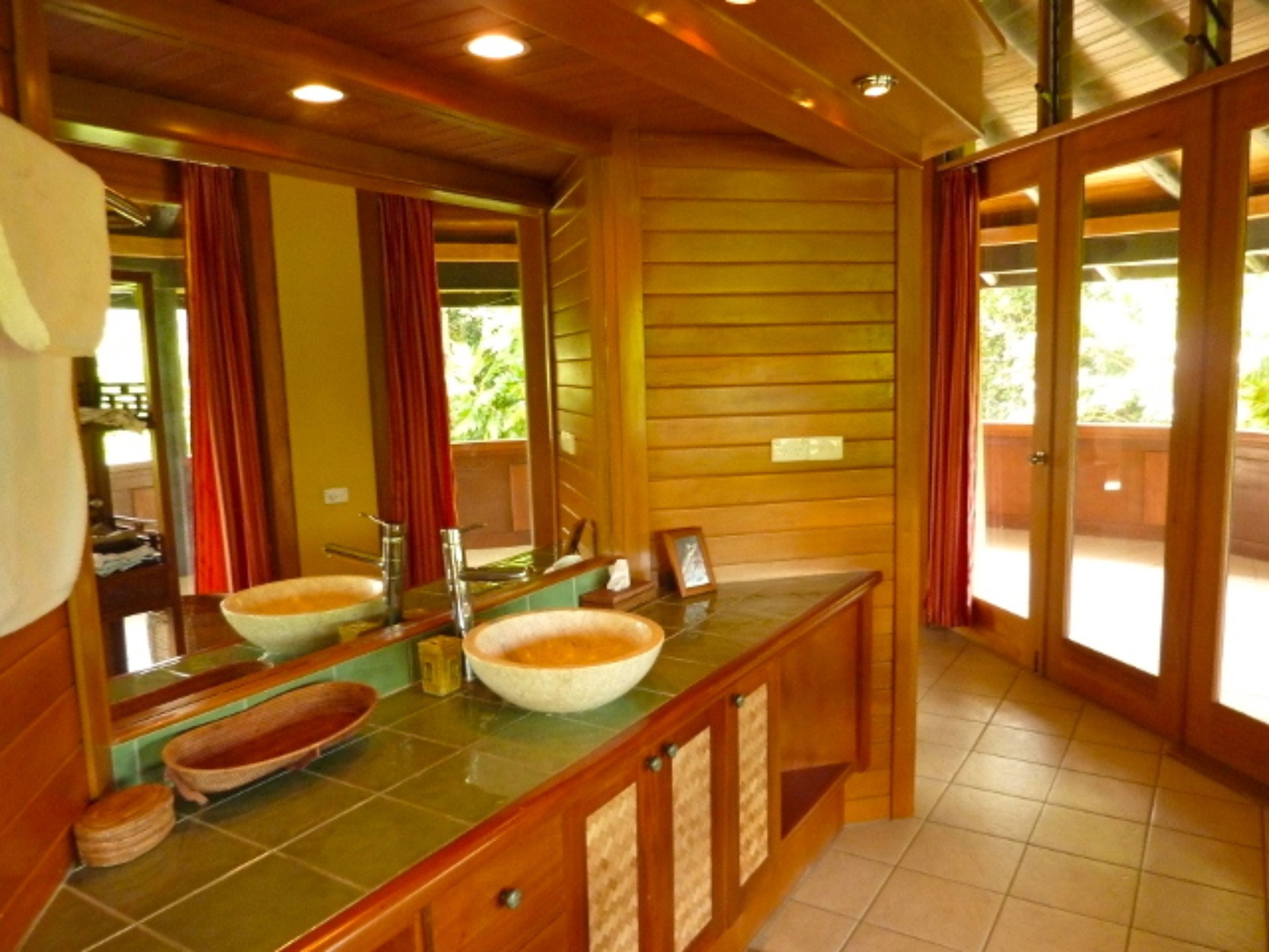 Een badkamer met houten lambrisering, twee wastafels met grote kommen, een spiegel en uitzicht op de buitenlucht door een glazen deur.