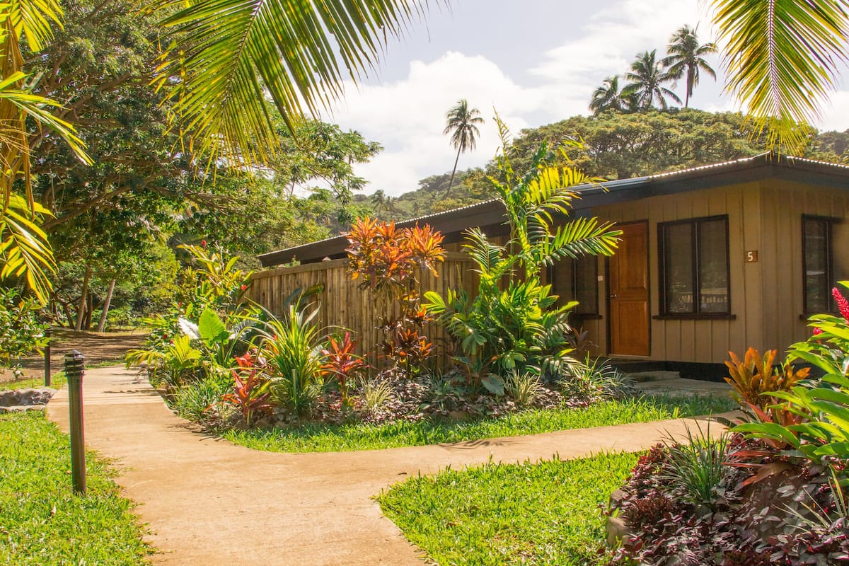 A bungalow with a wooden exterior, surrounded by lush greenery and a pathway leading to the entrance.