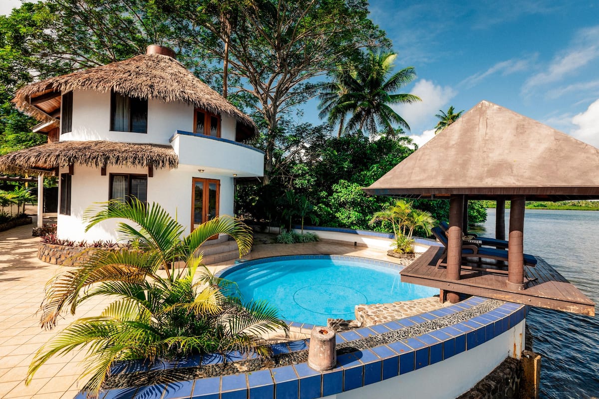 A two-story house with a thatched roof, a swimming pool, and a wooden deck overlooking a body of water.