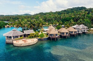An overwater bungalow resort with thatched-roof structures, clear blue water, and lush greenery in the background.