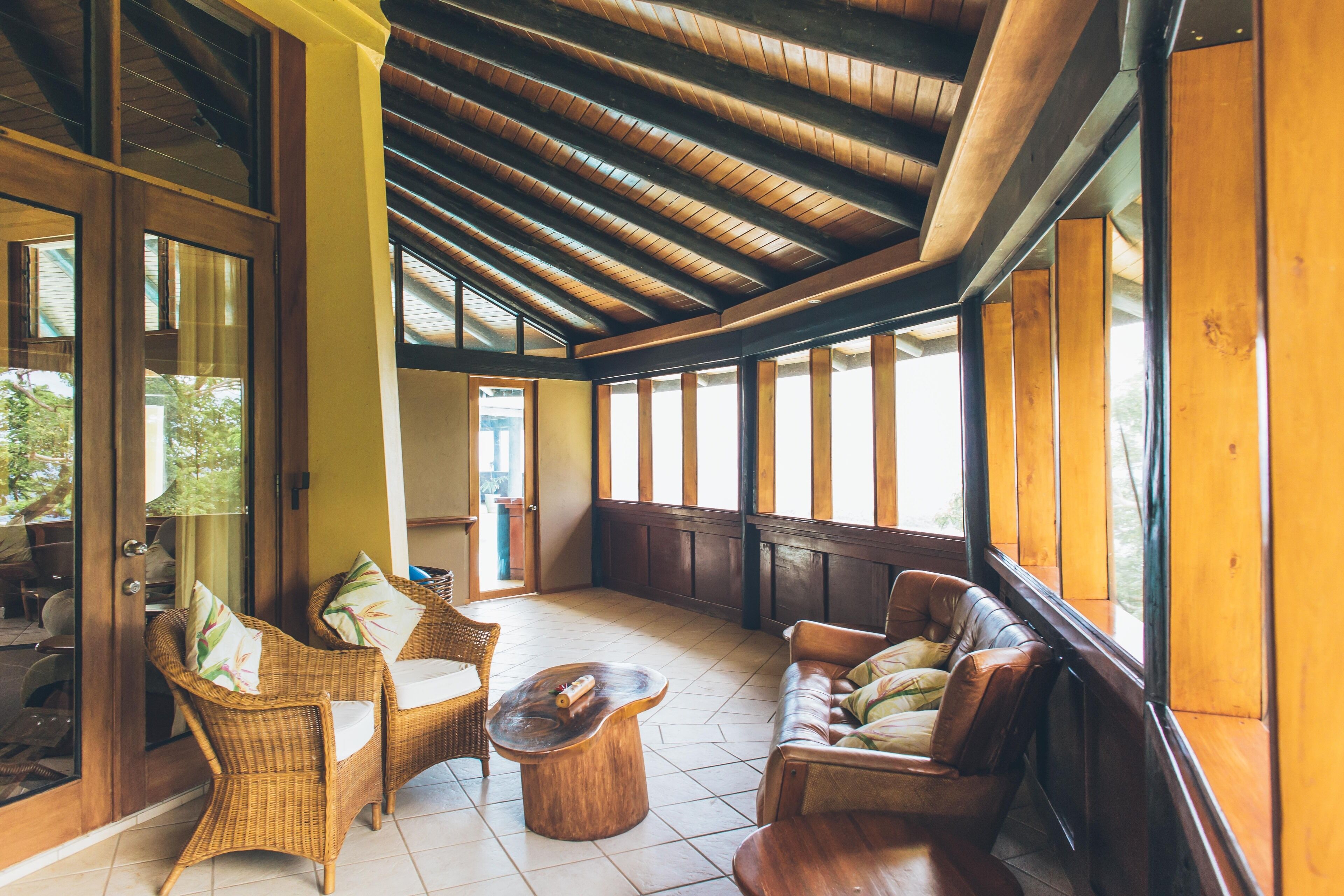A spacious indoor area with wooden ceiling beams, wicker furniture, and large windows providing natural light.