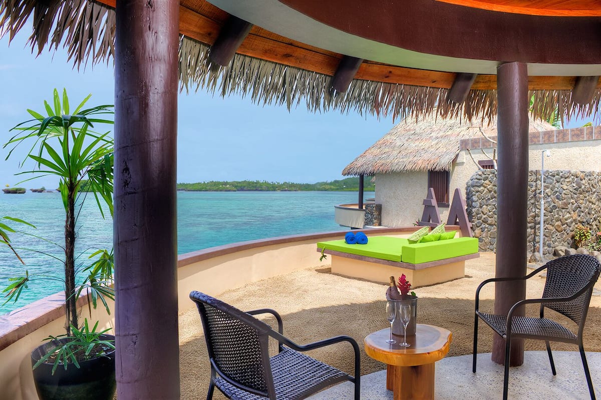 A tropical resort with a thatched-roof structure, a pool area with lounge chairs, and a view of the ocean.