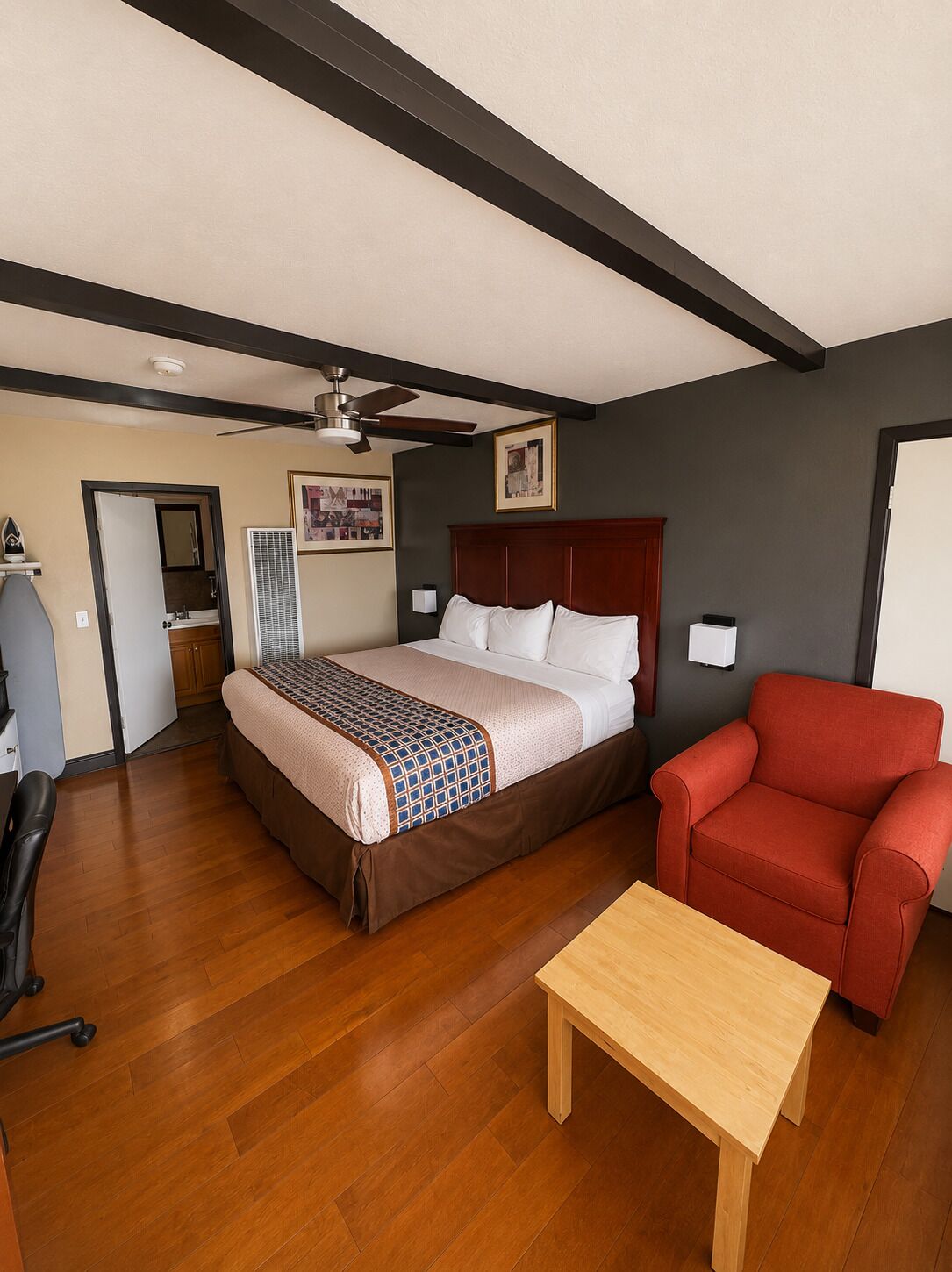 A hotel room with a bed, a red armchair, a wooden table, a ceiling fan, and a framed picture on the wall.